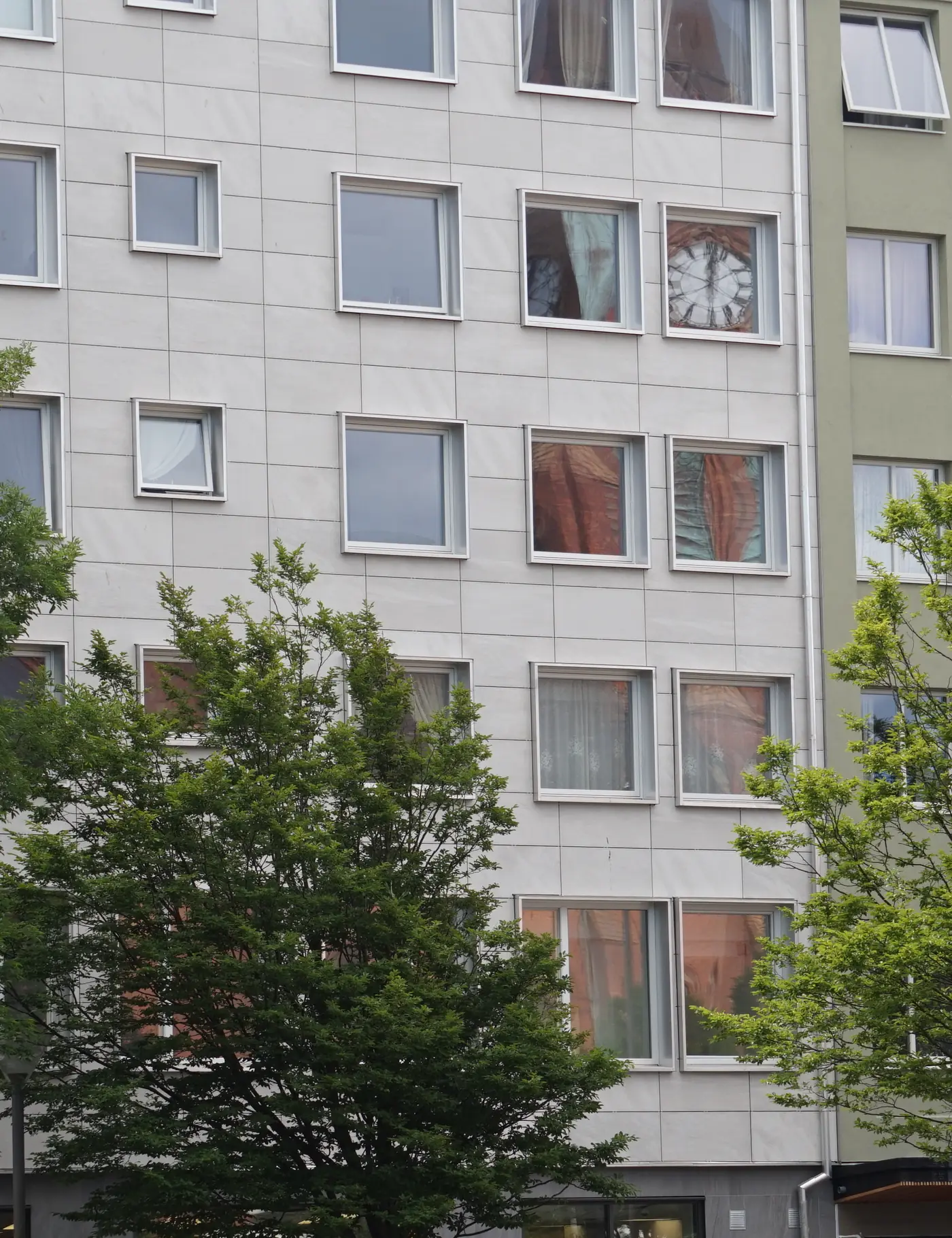 Ett flerbostadshus i Helsingborg med gardiner i fönstren, gröna träd framför fasaden och butiker i bottenvåningen